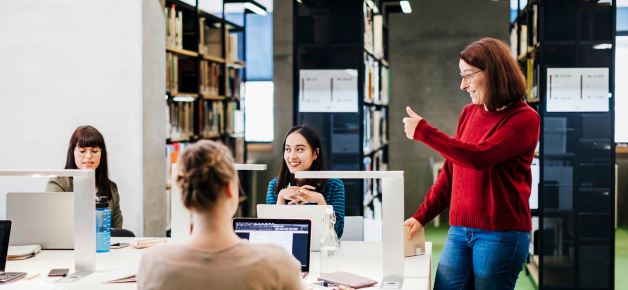 A woman standing up at the end of a large table giving a thumbs up to a seminar group.
