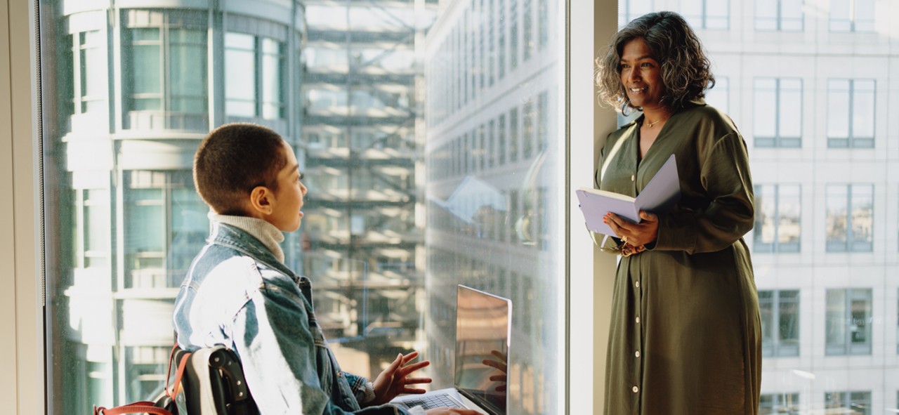 Businesswoman holding a notepad standing against tall office window talks to colleague