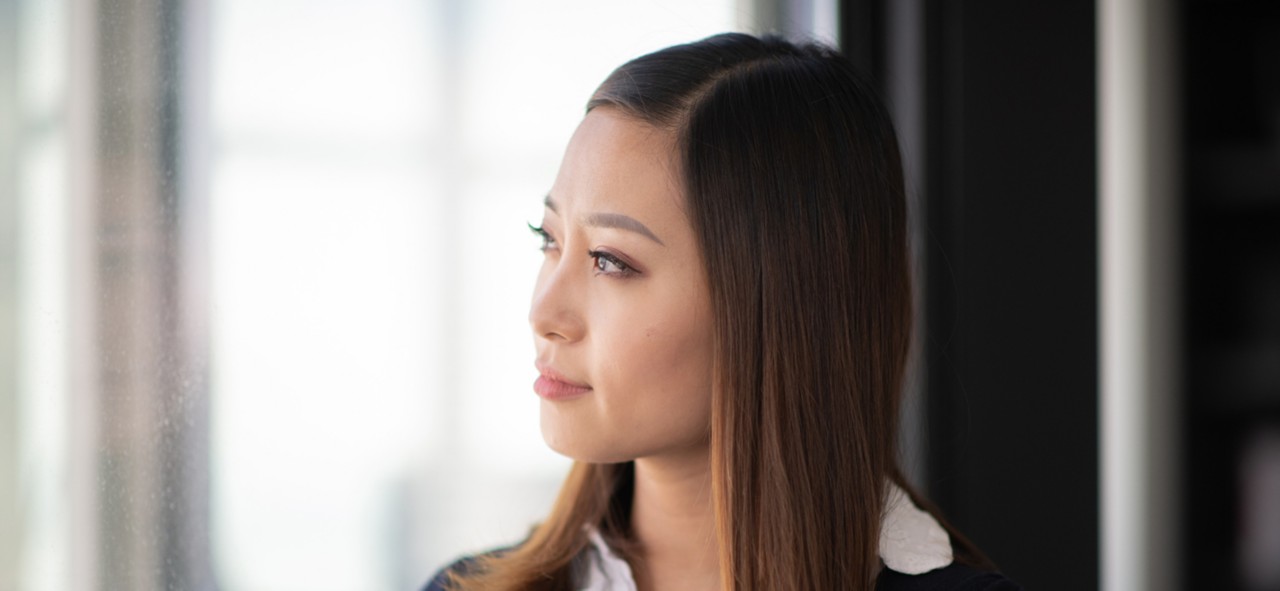 A beautiful Chinese woman is standing near a window indoors. She looks thoughtful and serious. The woman is holding a cup of tea. She is looking outside