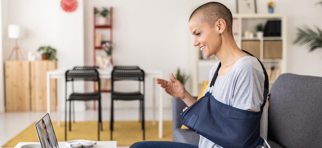 Woman in an arm sling using a laptop while having a video call with her doctor sitting on a sofa at home. Medicine and health care concept.